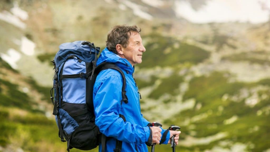 A smiling older man with a blue backpack and hiking poles looks to the right.