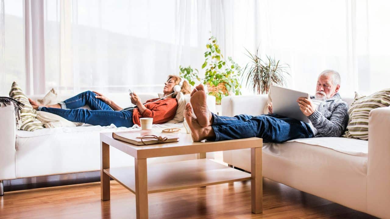 A man and a woman relax on separate couches, reading and using devices, in a bright living room.