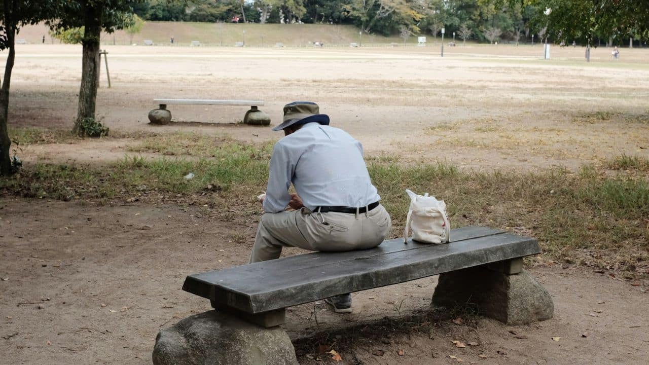 A man in a hat and light shirt sits alone on a park bench.