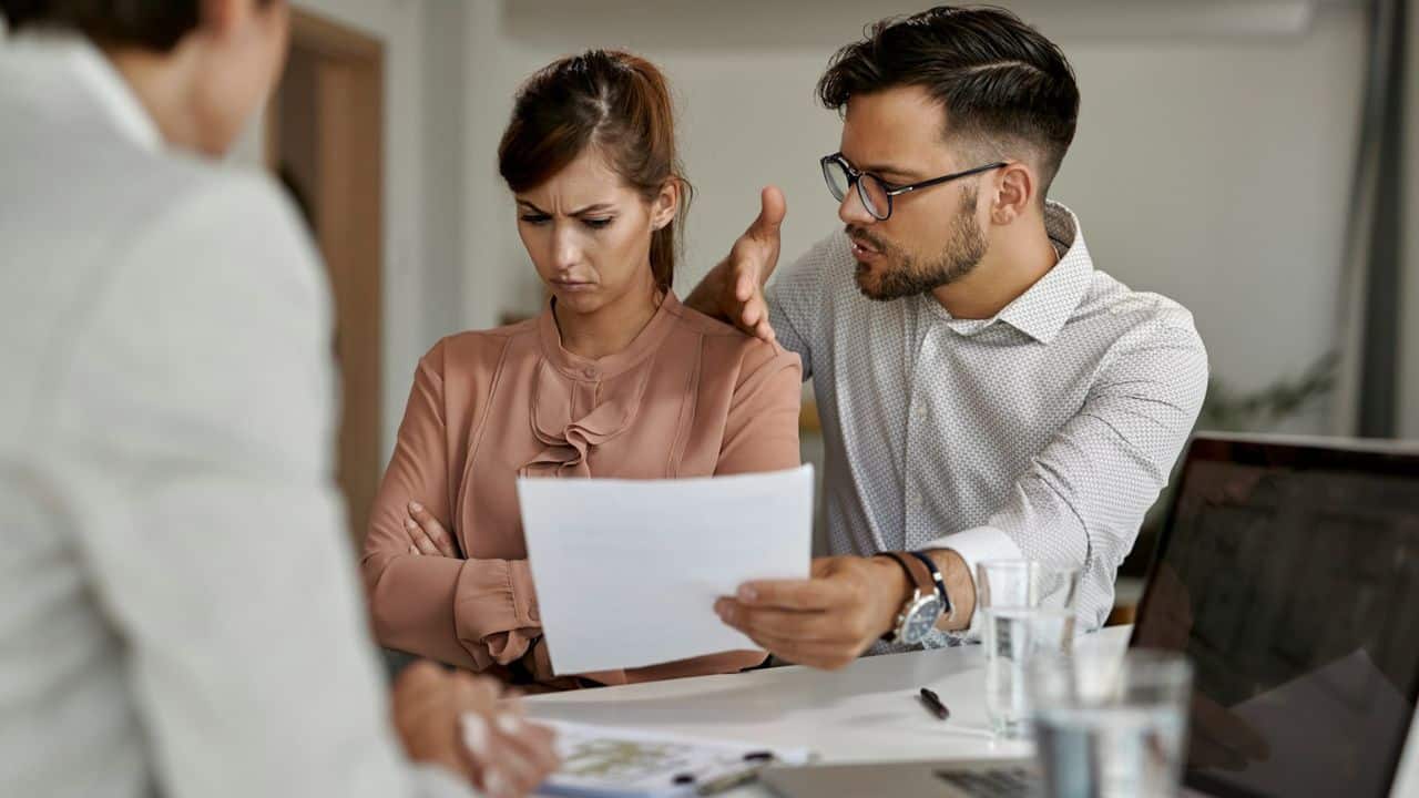 A man with glasses talks and gestures towards a woman with crossed arms, who looks displeased, while another person sits across from them.