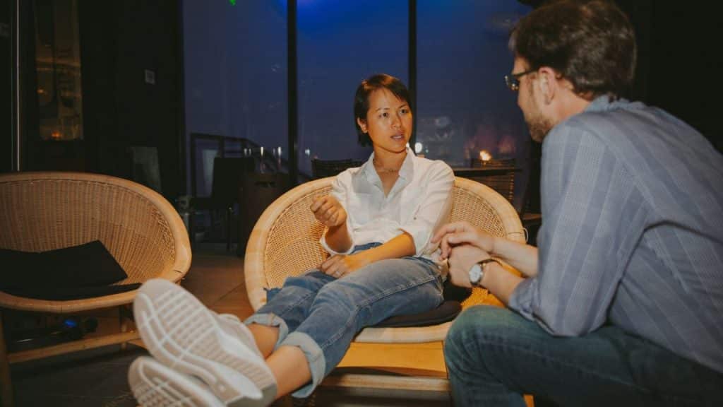 A woman in a white shirt and jeans sits in a wicker chair, talking to a man.