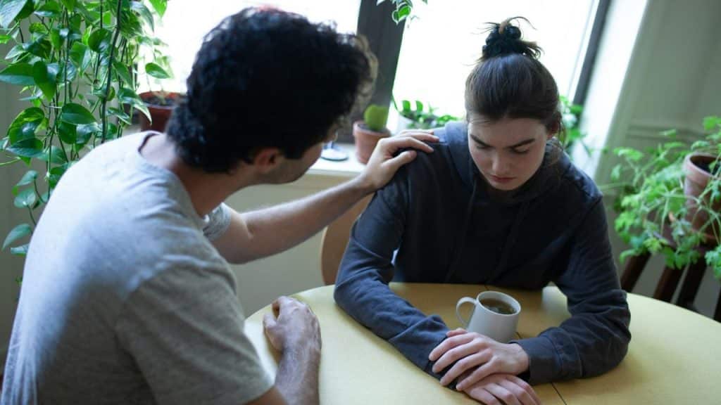 A man comforts a woman at a table with a mug, surrounded by plants.