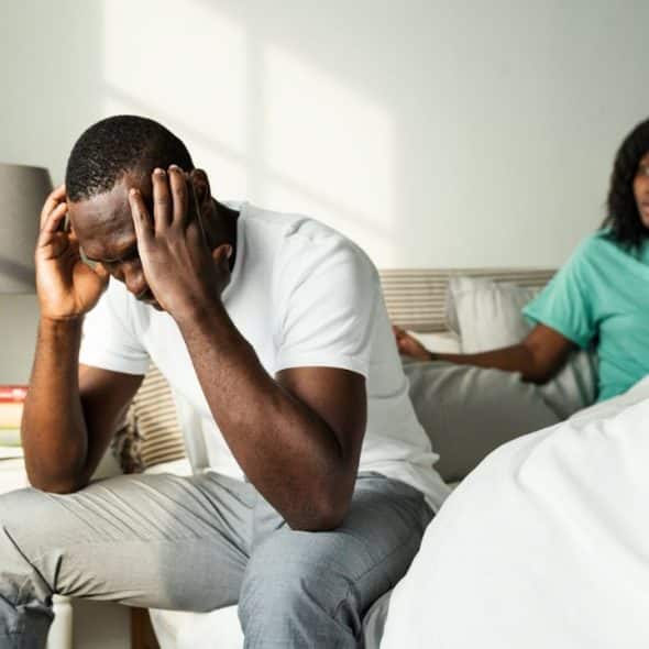 A man sits on a bed with his head in his hands, while a woman gestures in the background.