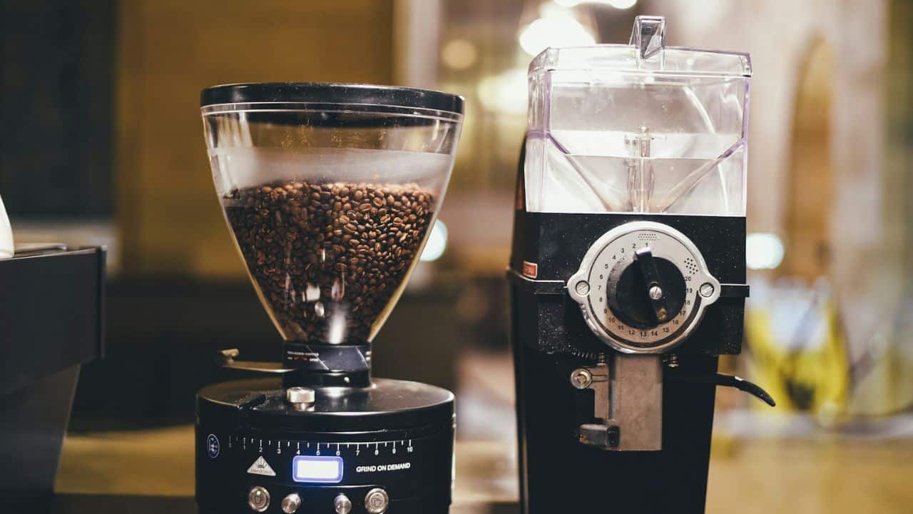 A clear hopper full of coffee beans sits atop a coffee grinder, next to another grinder.