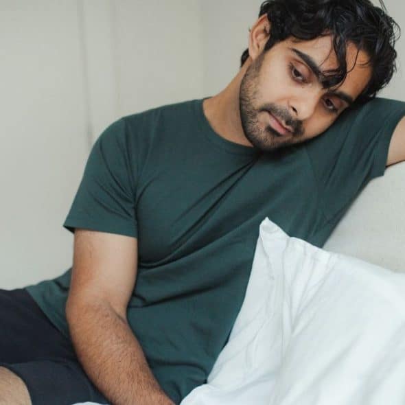 A man in a green shirt and black shorts sits on a bed, looking downcast.