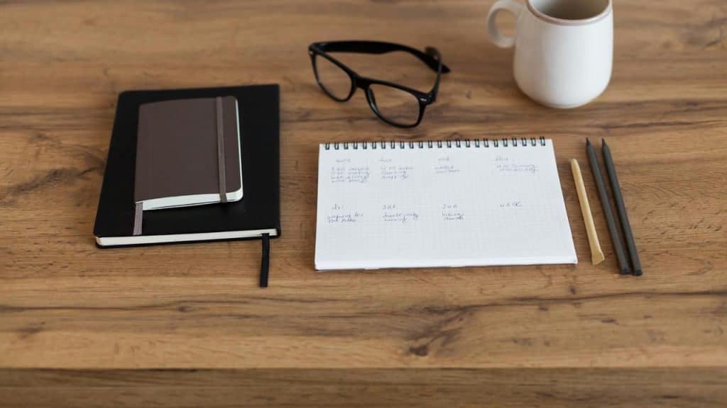 A wooden desk with notebooks, glasses, a planner, pens, and a mug.