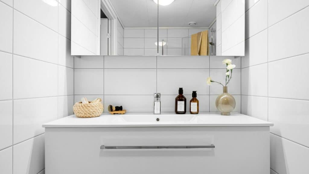 A minimalist bathroom with white tiles features a sink, mirror, and organized toiletries.