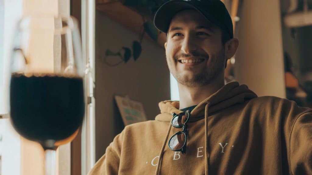 A man in a hoodie and cap smiling beside a dark drink in the foreground.