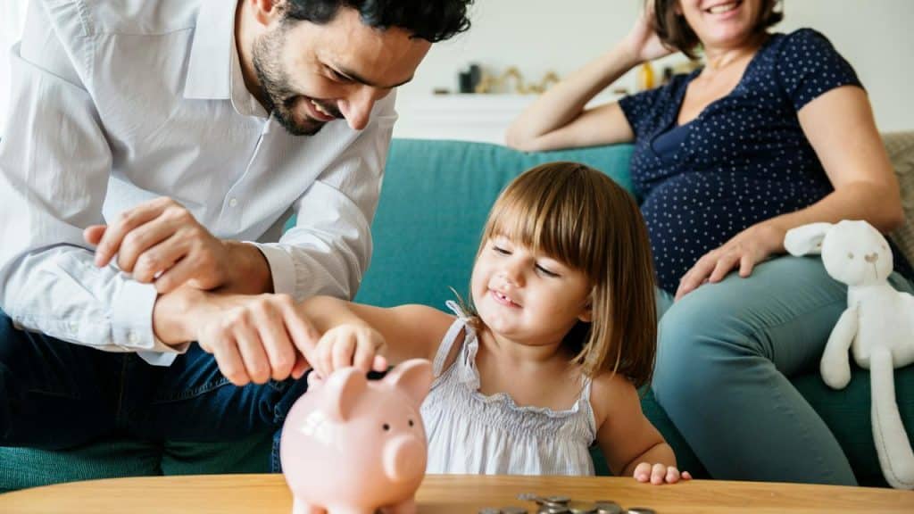 A man and girl adding a coin to a piggy bank while a pregnant woman watches.