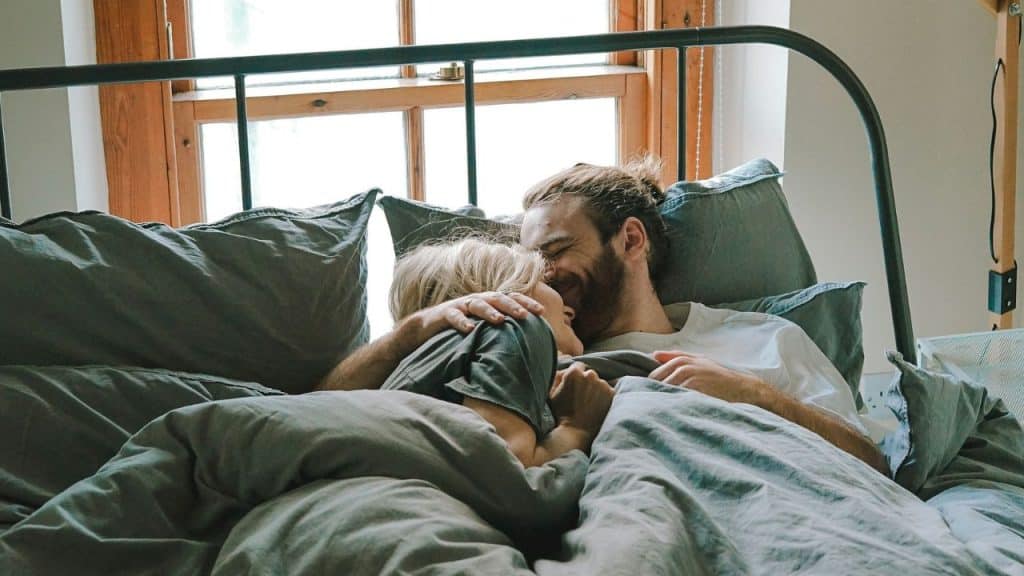 A couple smiles and cuddles together in bed under gray blankets.