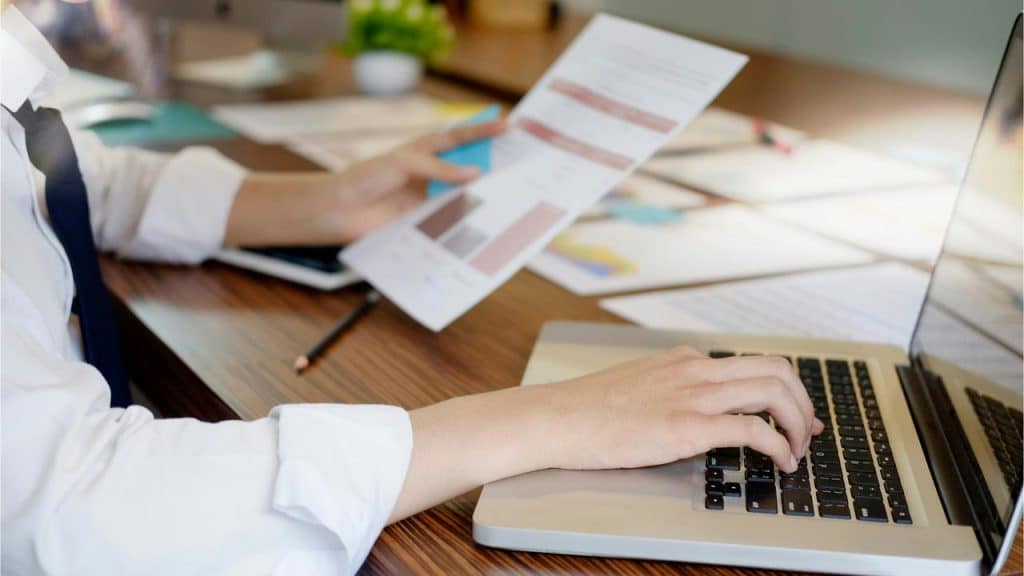A person holding a charted report while typing on a laptop.