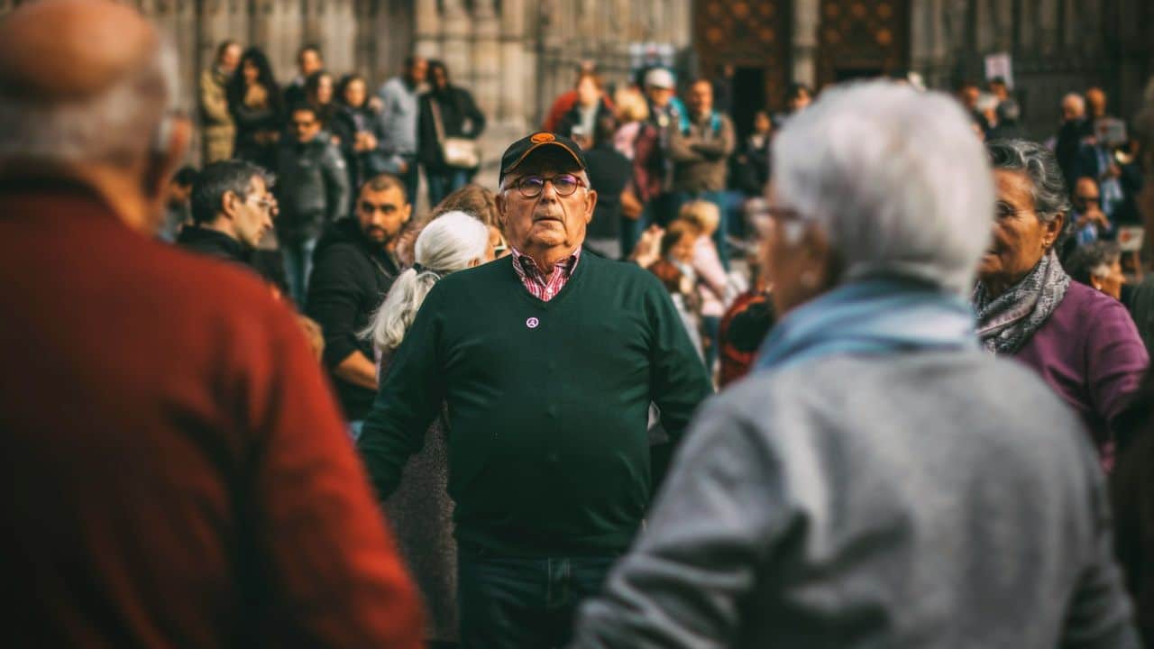 A man in a dark green sweater and cap looks directly at the camera in a crowd.