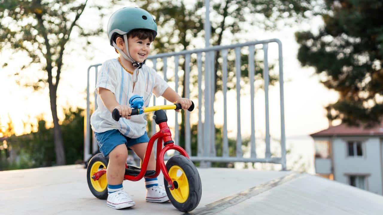 A toddler wearing a helmet riding a small red balance bike outdoors.