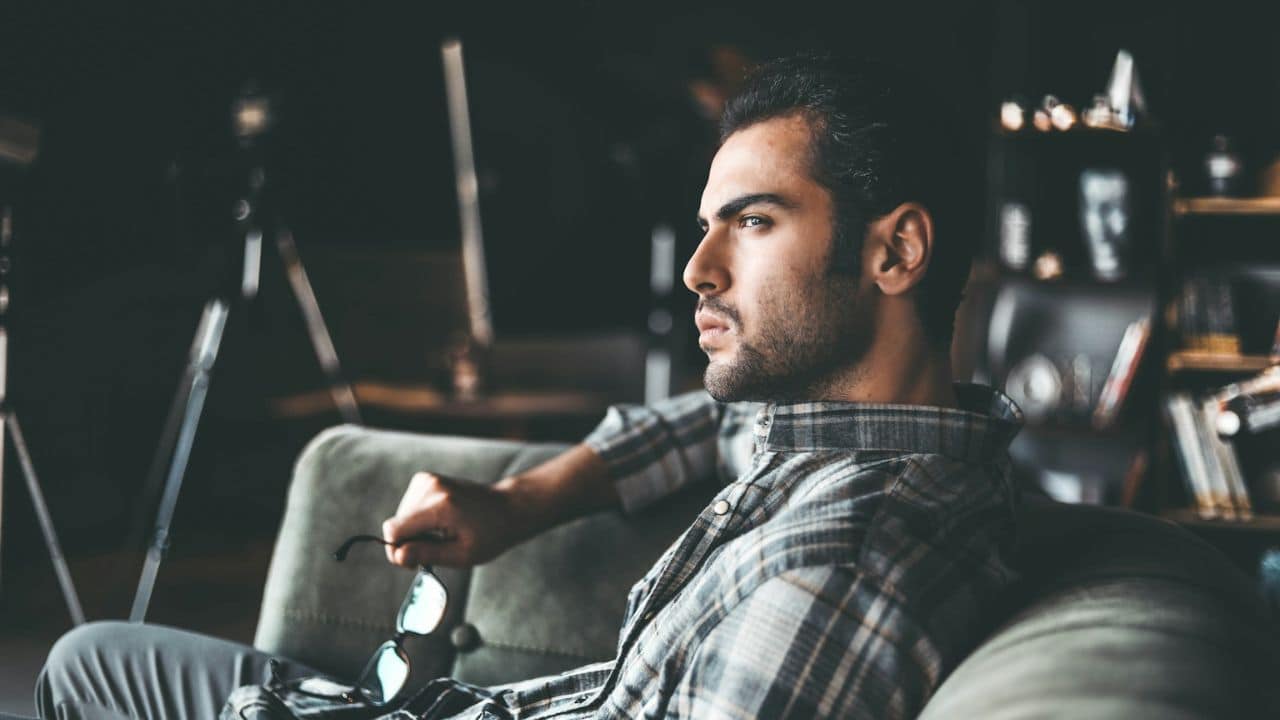 A man with dark hair and a beard, wearing a plaid shirt, sits on a couch holding glasses, looking away.