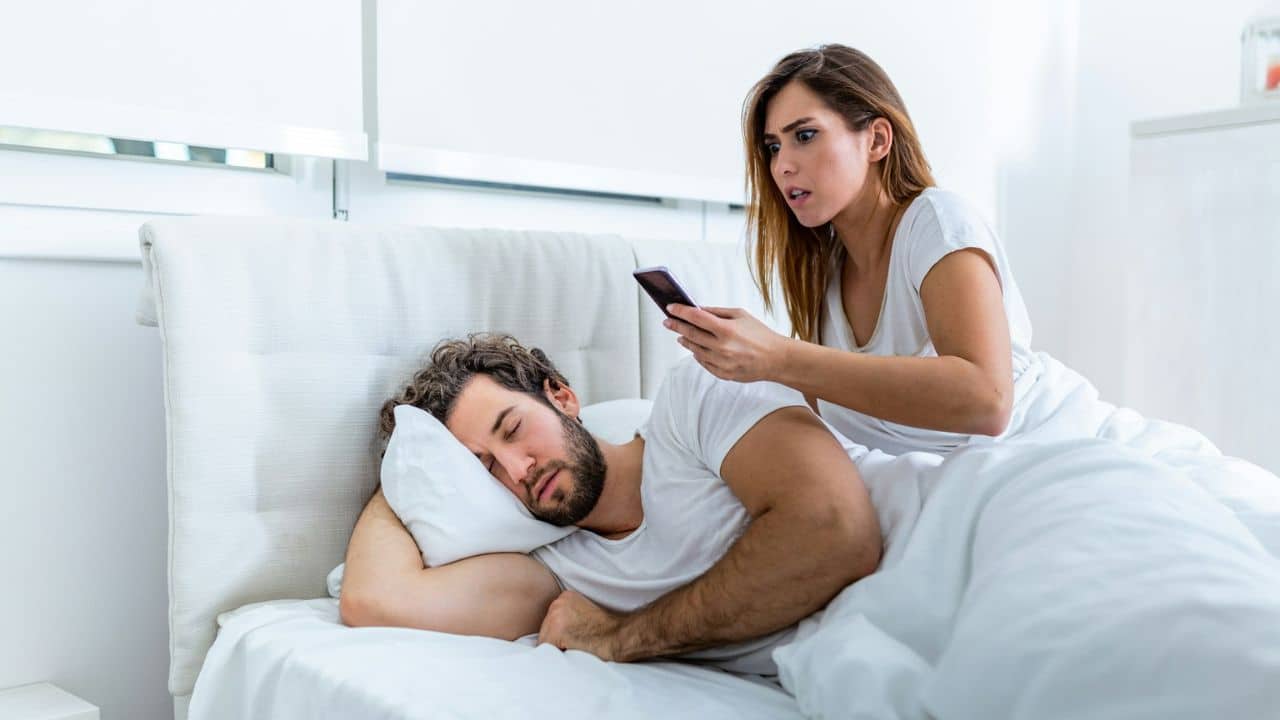 A woman in bed looks shocked while holding a phone over a sleeping man.