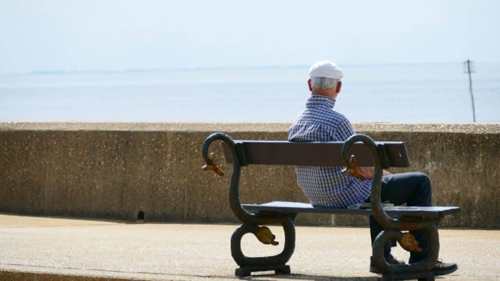 A man in a plaid shirt and white cap sits on a decorative bench facing the ocean.