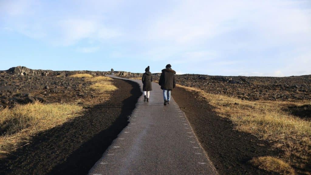 A view from behind two people walking on a paved path through a rocky, grassy landscape.