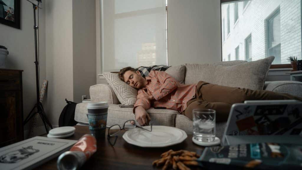 A man asleep on a sofa beside a cluttered coffee table.