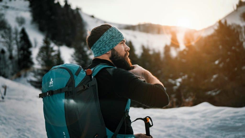 A bearded hiker with a backpack gazing at a snowy mountain sunset.