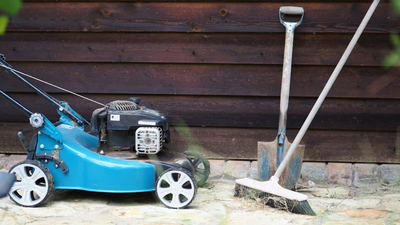 A blue lawnmower, a shovel, and a broom lean against a wooden fence.