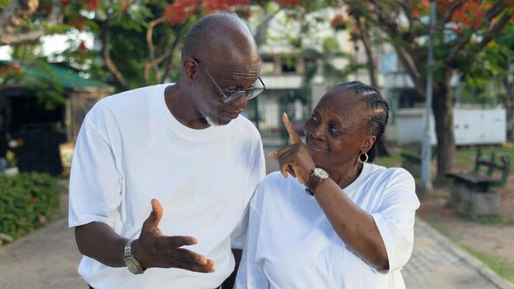 A smiling older Black couple in white shirts interacts playfully outdoors.