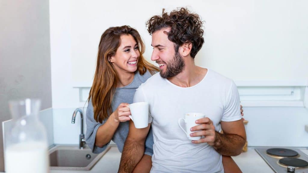 A couple sharing coffee mugs in a bright kitchen.