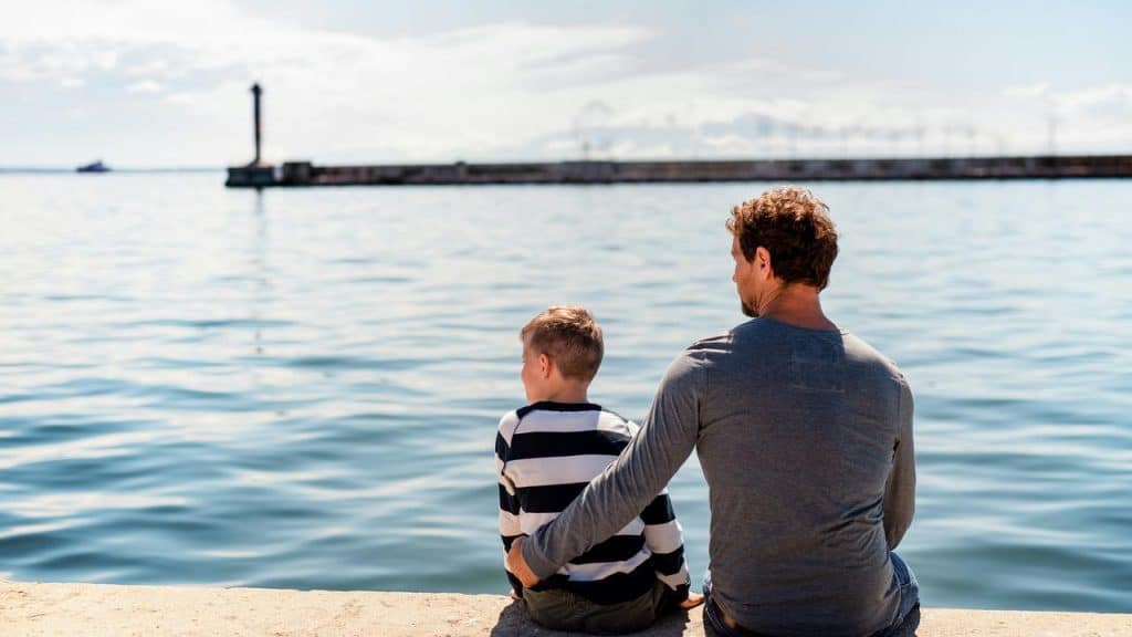 A man and a young boy sitting on a pier, gazing over calm water.