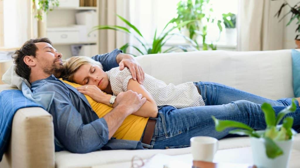 A man and woman are sleeping and cuddling on a couch in a living room.