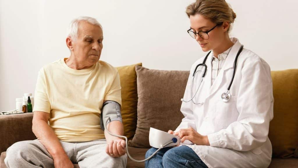 A healthcare professional takes an elderly man's blood pressure while he sits on a couch.
