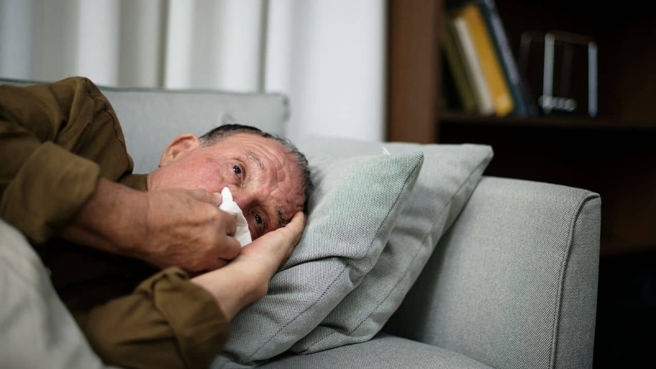 A man lies on a sofa, wiping his nose with a tissue.