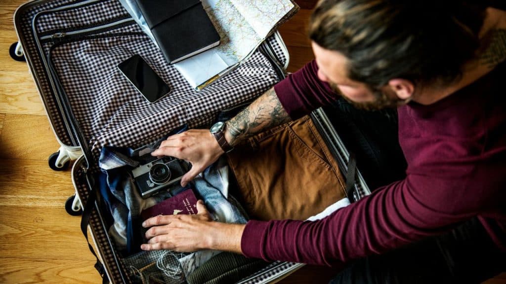A man with tattooed arms packs a suitcase with clothes, a camera, and a passport.