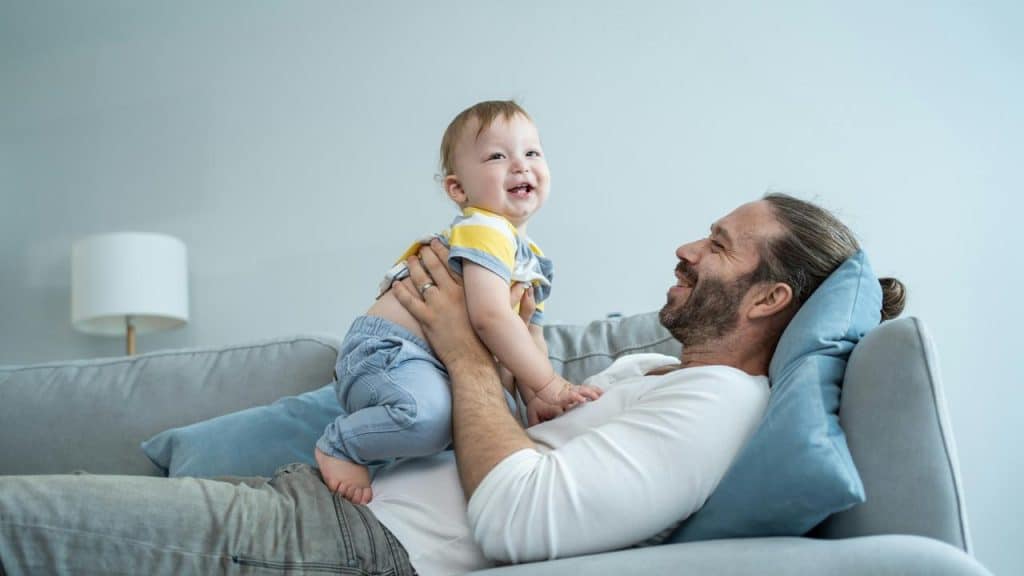 A man lifting a smiling baby on a couch.