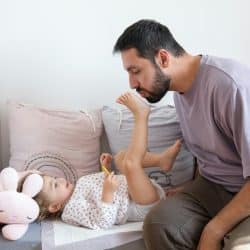 A man kissing a child’s foot as she lies on a cushion beside a stuffed bunny.