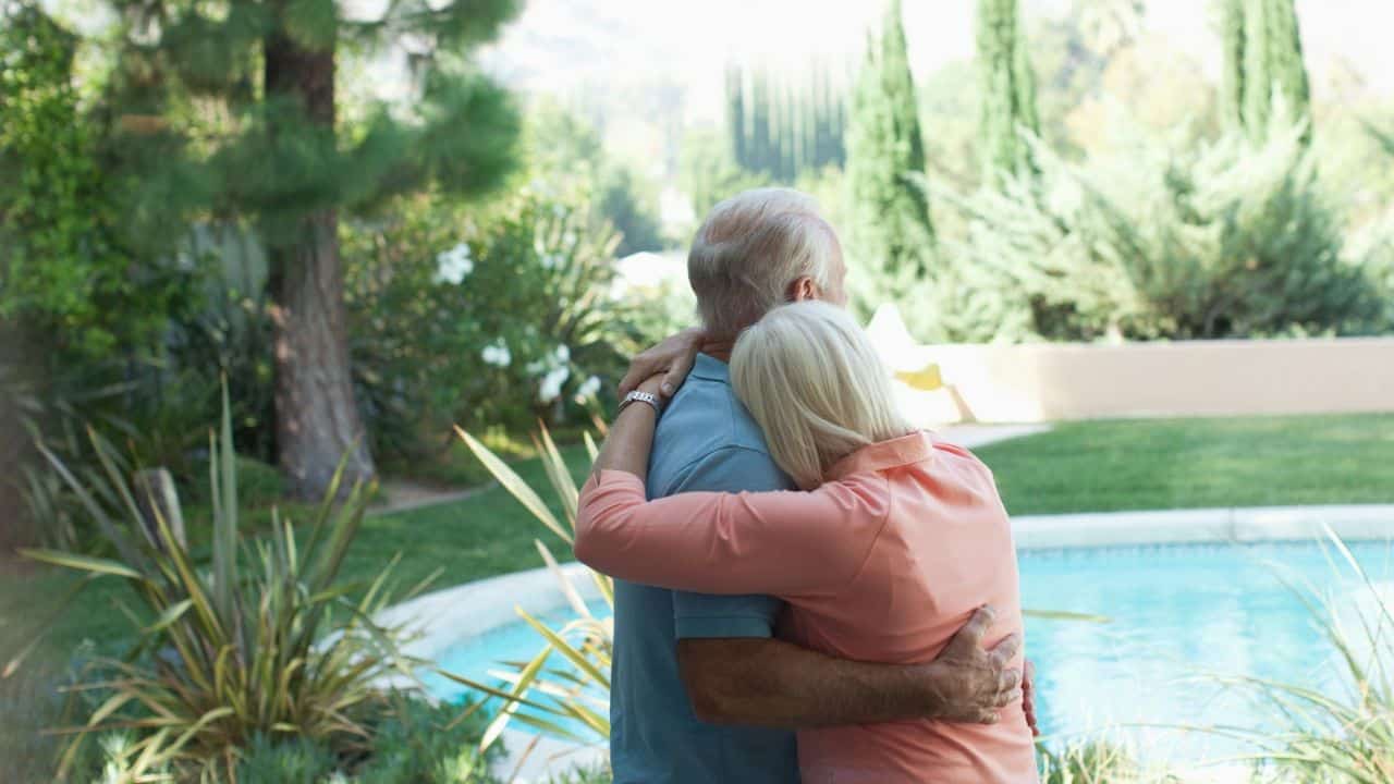A couple with gray hair embraces outdoors by a swimming pool.