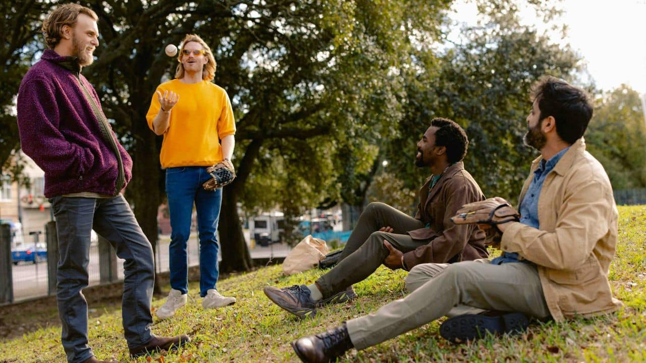 Four men are outdoors; one throws a baseball, two sit on grass, and one stands.