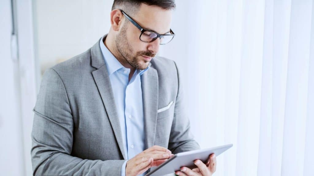 A man in glasses and a suit looks at a tablet.