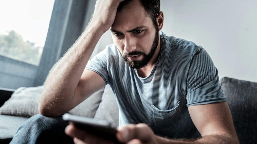 A distraught bearded man with his hand on his head looks at his phone.