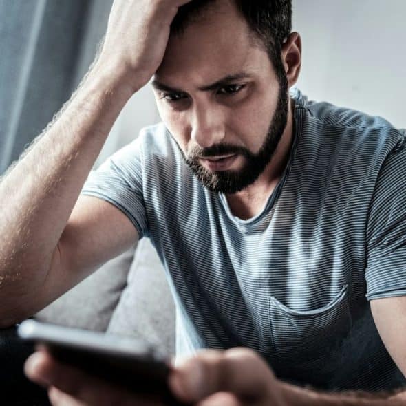 A distraught bearded man with his hand on his head looks at his phone.