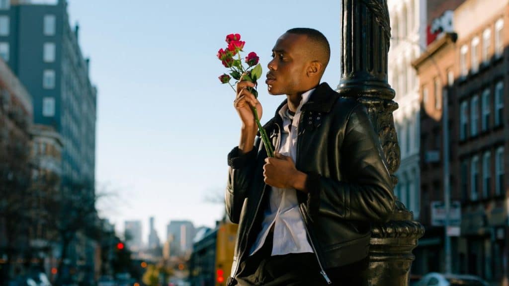 A man in a leather jacket smells a bouquet of red roses on a city street.
