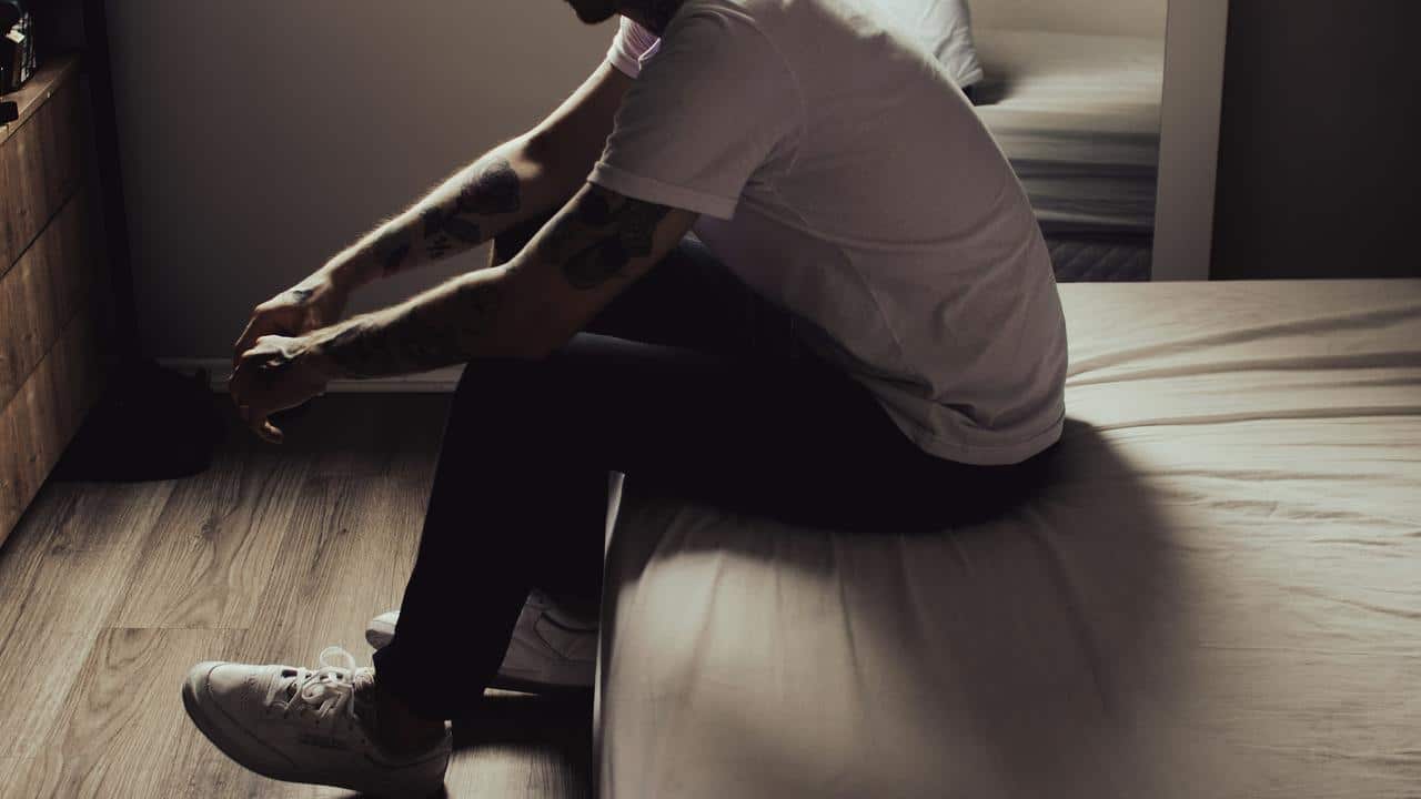 A tattooed man sitting on the edge of a bed looking down.