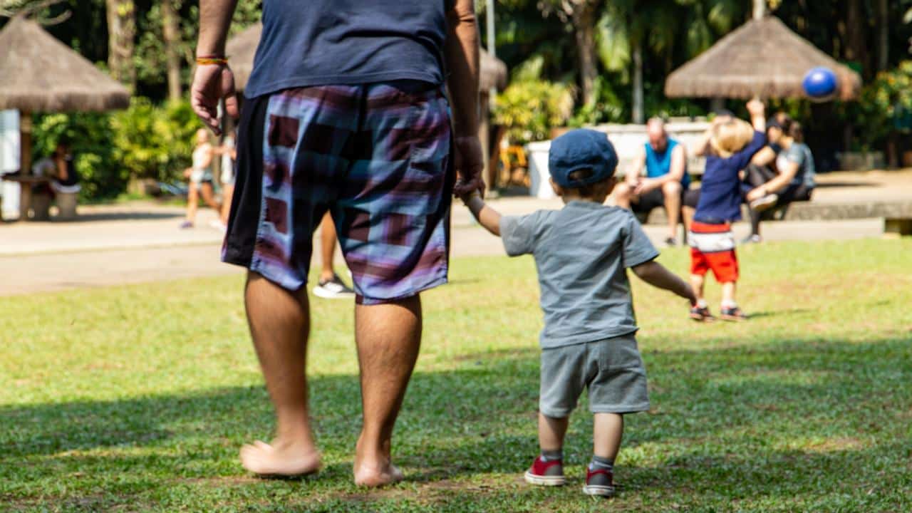 A man holding a toddlerβs hand while walking barefoot in a park.