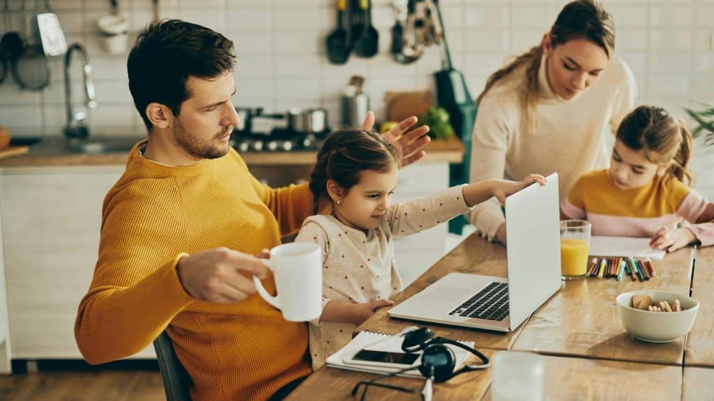 A father at a kitchen table with his daughters using a laptop and drawing.