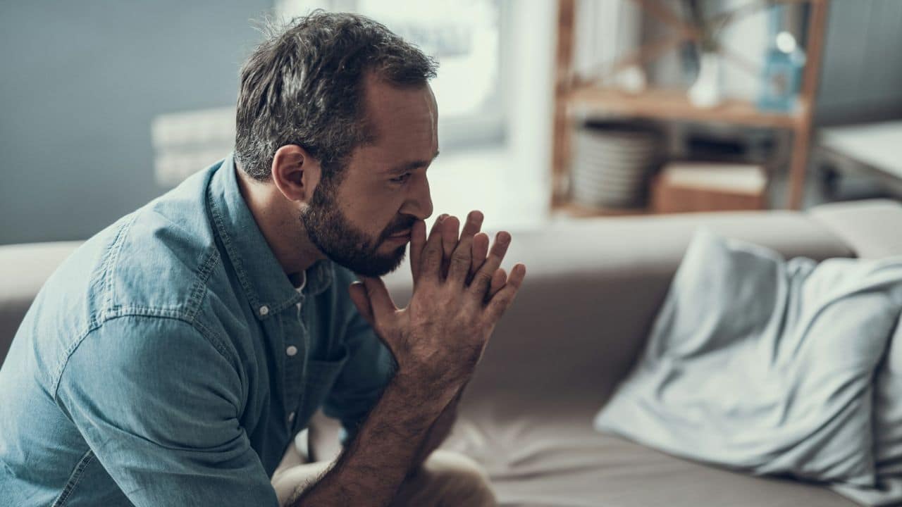 A thoughtful man with a beard sits on a couch with his hands clasped under his chin.
