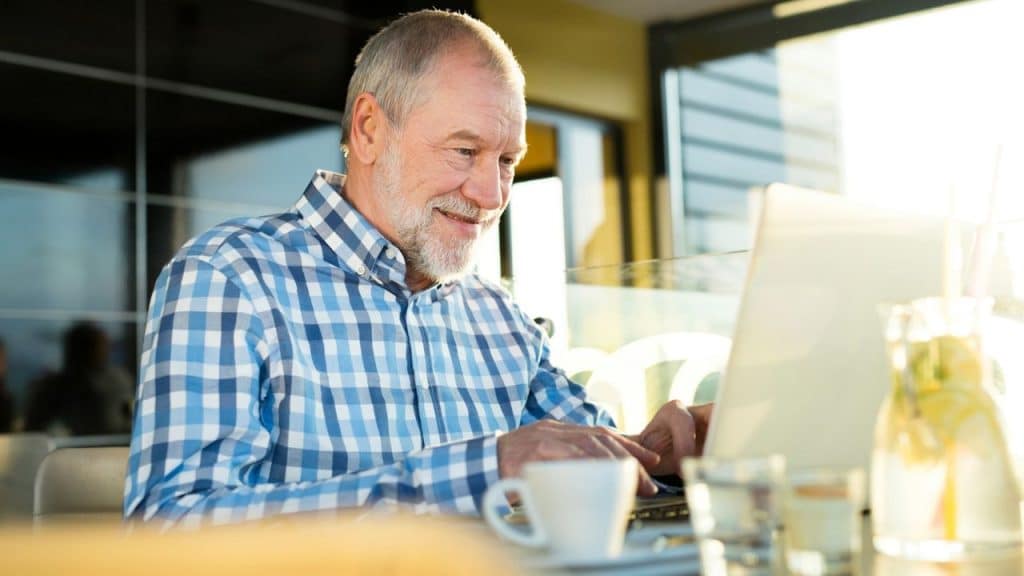 A smiling older man with a beard sits at a table, looking at a laptop with a cup and glasses nearby.