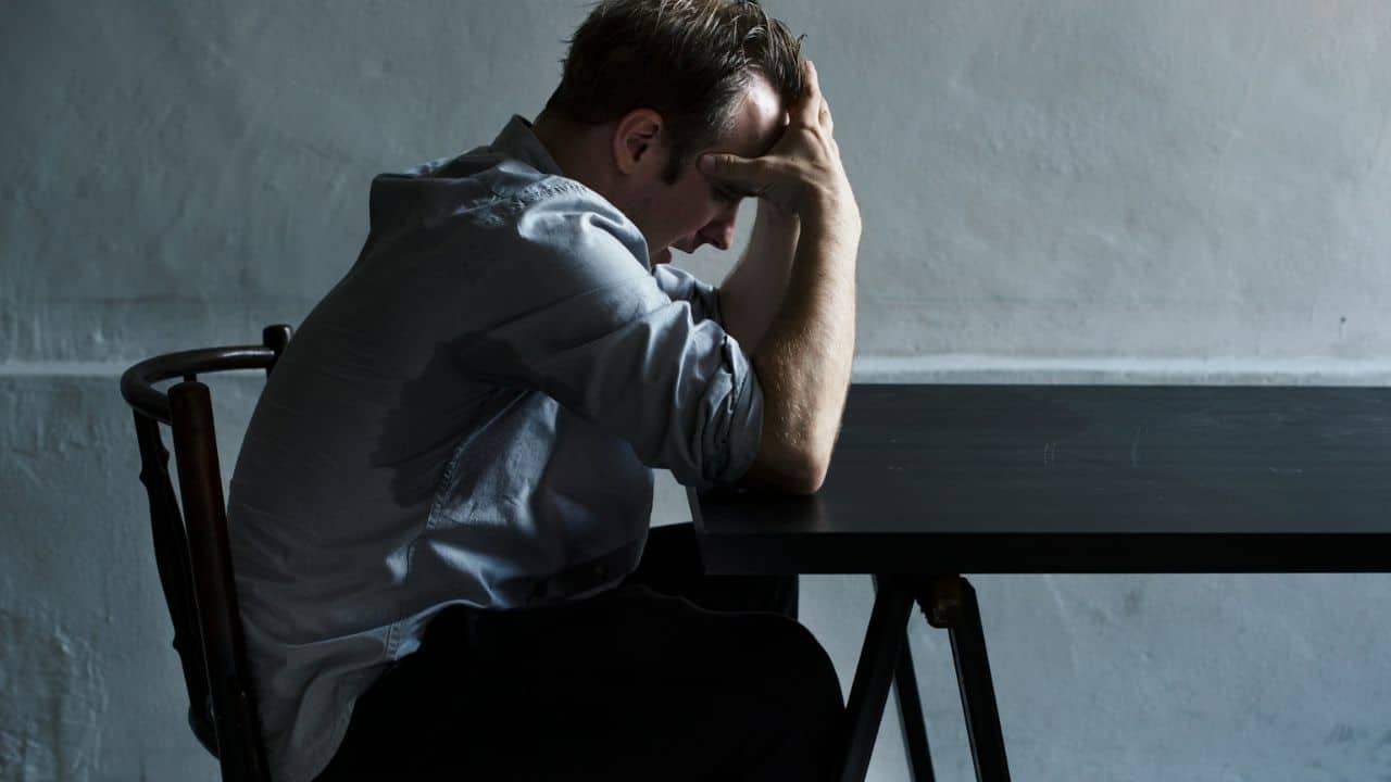 A man in a light blue shirt sits at a dark table, with his head in his hands.