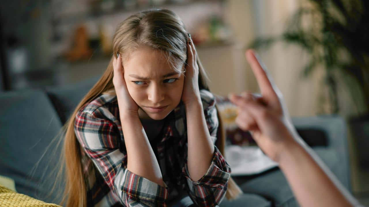 A young girl covers her ears and looks away from a pointing finger.