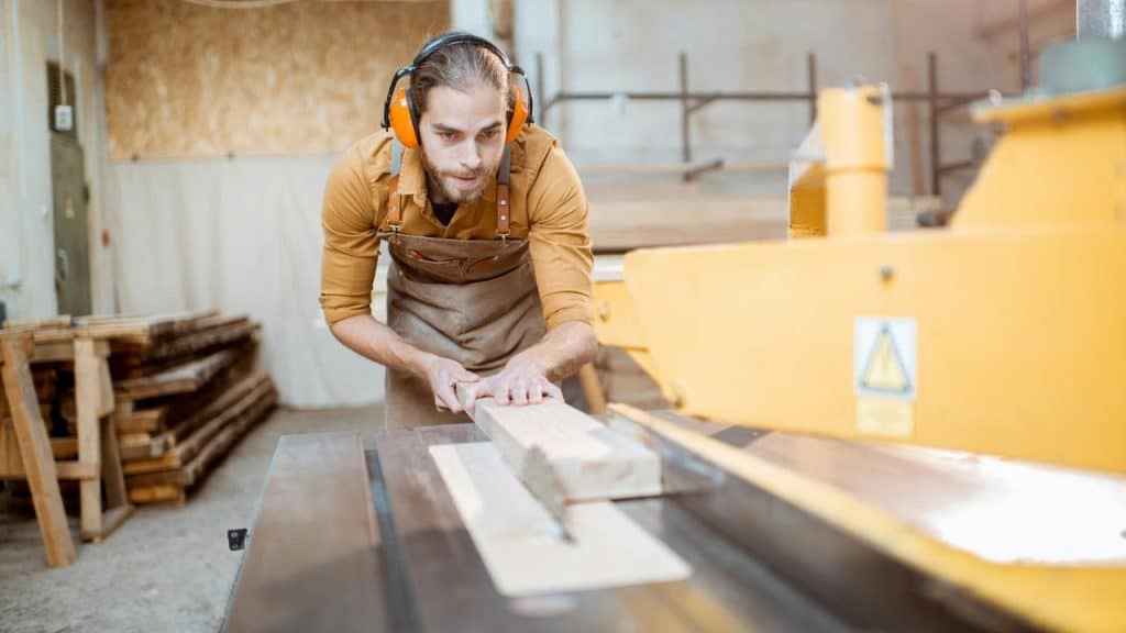 A woodworker with ear protection pushing a wooden board through a jointer planer machine.
