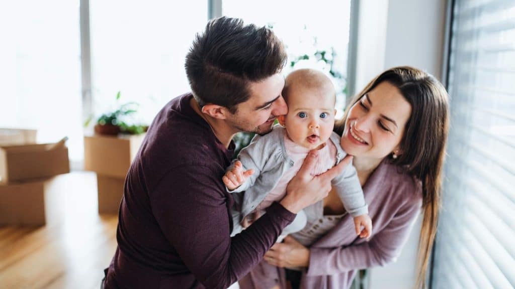 A couple playing with their wide‑eyed baby in a bright room with moving boxes.