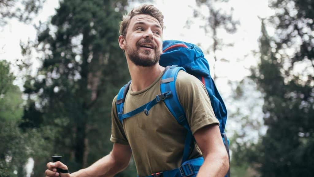 A smiling man with a beard and backpack holds a hiking pole.