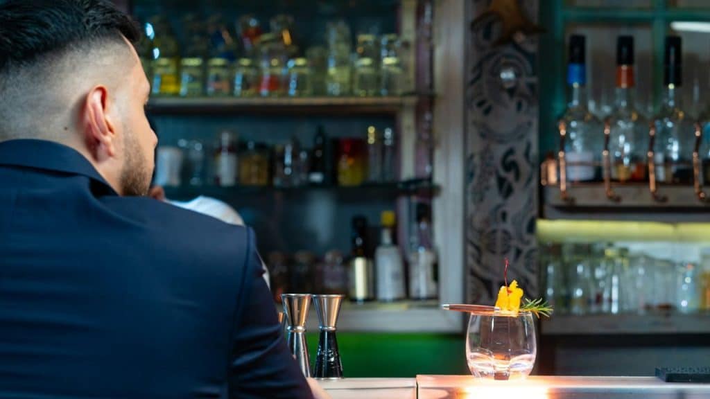 A person with a sharp haircut sits at a bar, looking at a cocktail.